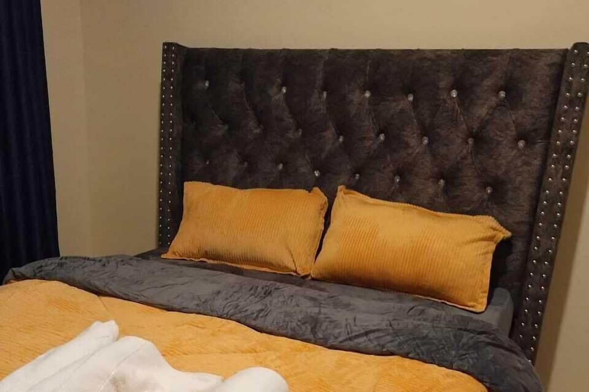 Un lit doté d’une tête de lit capitonnée, de deux coussins de couleur moutarde et d’une serviette blanche roulée posée sur un couvre-lit orange à texture.