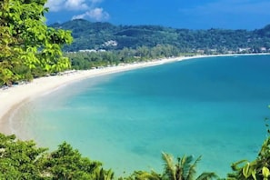 A beach with clear turquoise water, a sandy shore, and lush greenery in the background. - Panoramic sea-view condo near Kamala Beach (Kamala)