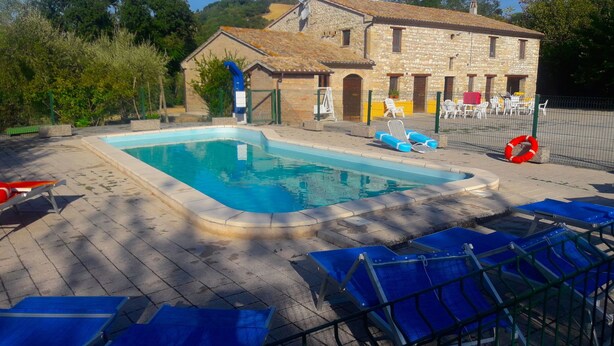 A swimming pool with blue lounge chairs, a stone building with a tiled roof, and a fenced area with chairs and a lifebuoy.