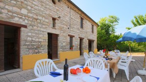 An outdoor dining area with stone walls, white plastic chairs, and a table set with a bottle, fruit, and flowers.
