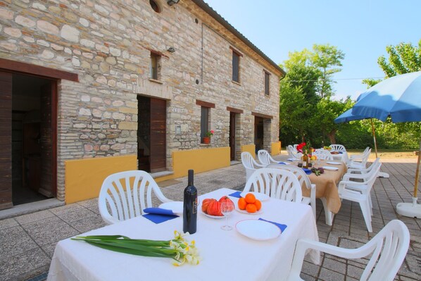 An outdoor dining area with stone walls, white plastic chairs, and a table set with a bottle, fruit, and flowers.
