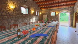 A dining area with a long table set for a meal, stone walls, wooden beams, and a view through an open archway.