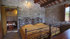 A bedroom with a stone wall, a bed with a metal frame, a wooden table with a chair, and a window with a view.