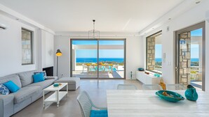 A modern living room with a grey sofa, white coffee table, and blue cushioned chairs. A large sliding glass door leads to an outdoor terrace with a pool and dining area. - Villa in Rhodes with Infinity Pool (Kalythies)