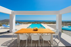 Ett träbord med vita flätade stolar, en pool och utsikt över havet.