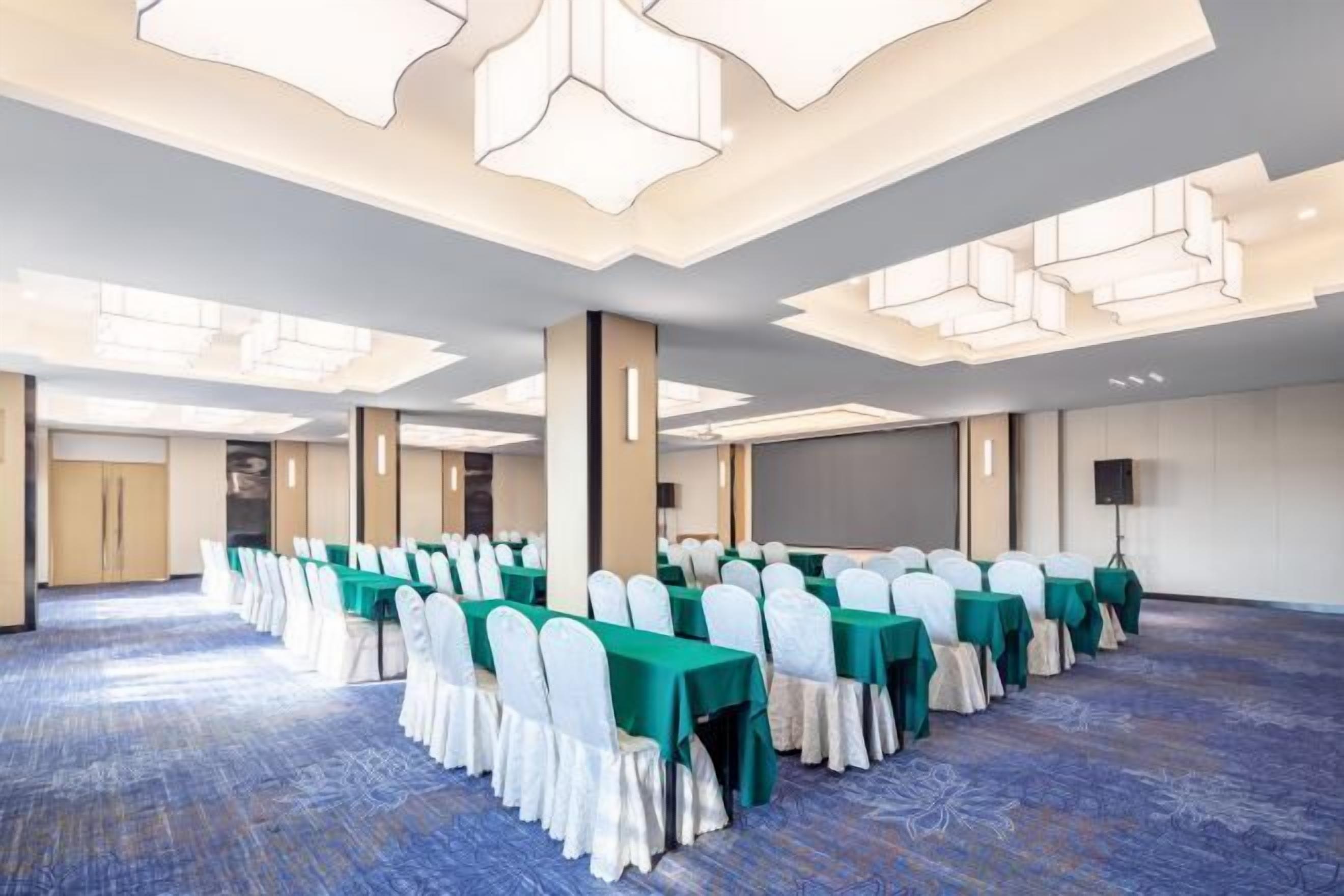 Un salón de conferencias con filas de mesas y sillas, suelo alfombrado y un diseño moderno en el techo.