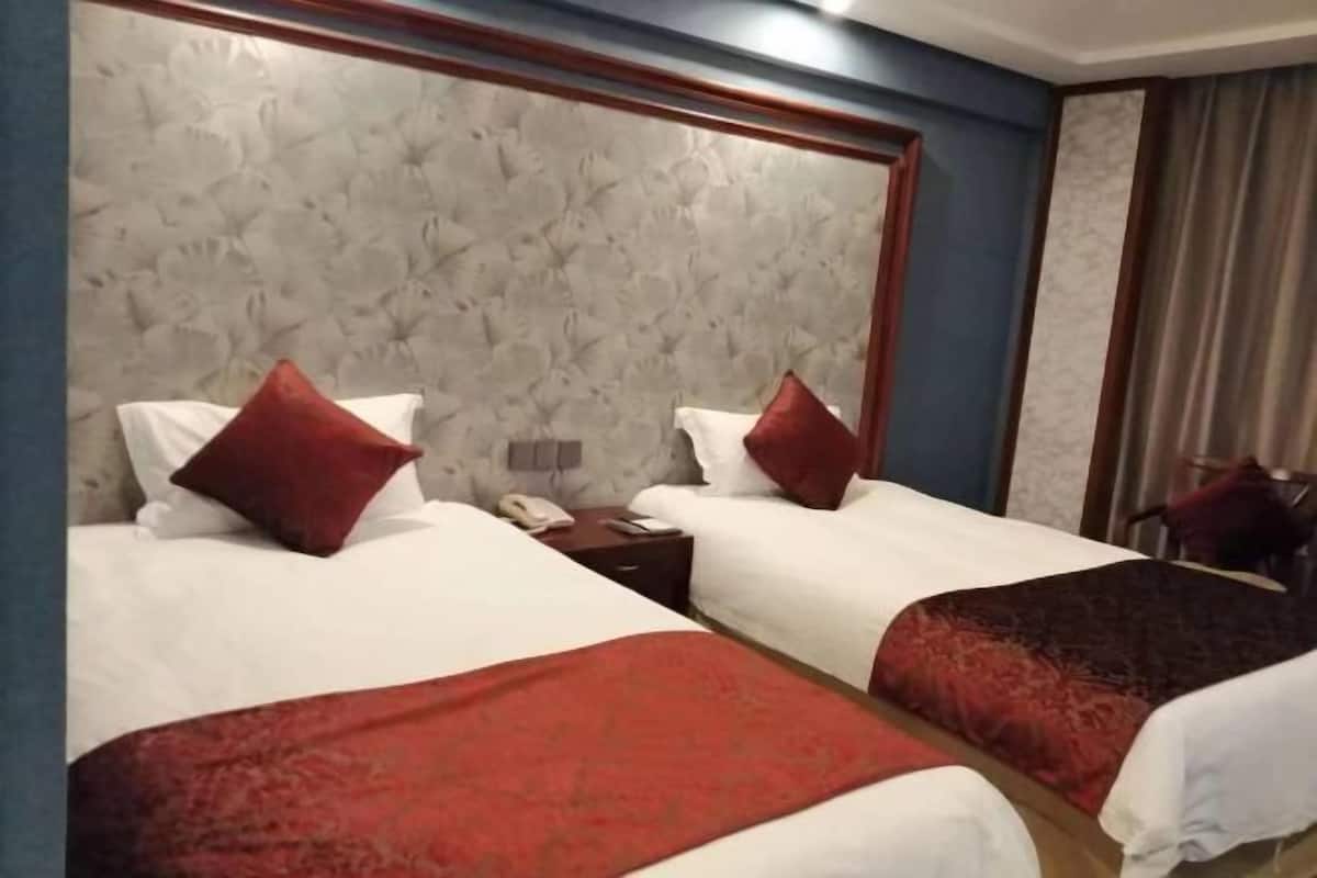 Habitación de hotel con dos camas, cada una con sábanas blancas y almohadas rojas, un cabecero de madera, una mesita de noche con teléfono y una ventana con cortinas.