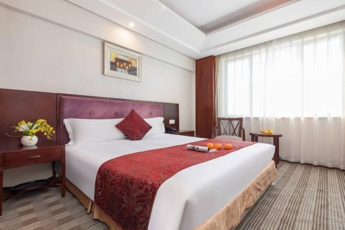 Habitación de hotel con una cama grande, cabecera de madera, mesita de noche con un florero, una pequeña mesa con fruta y una ventana con cortinas.