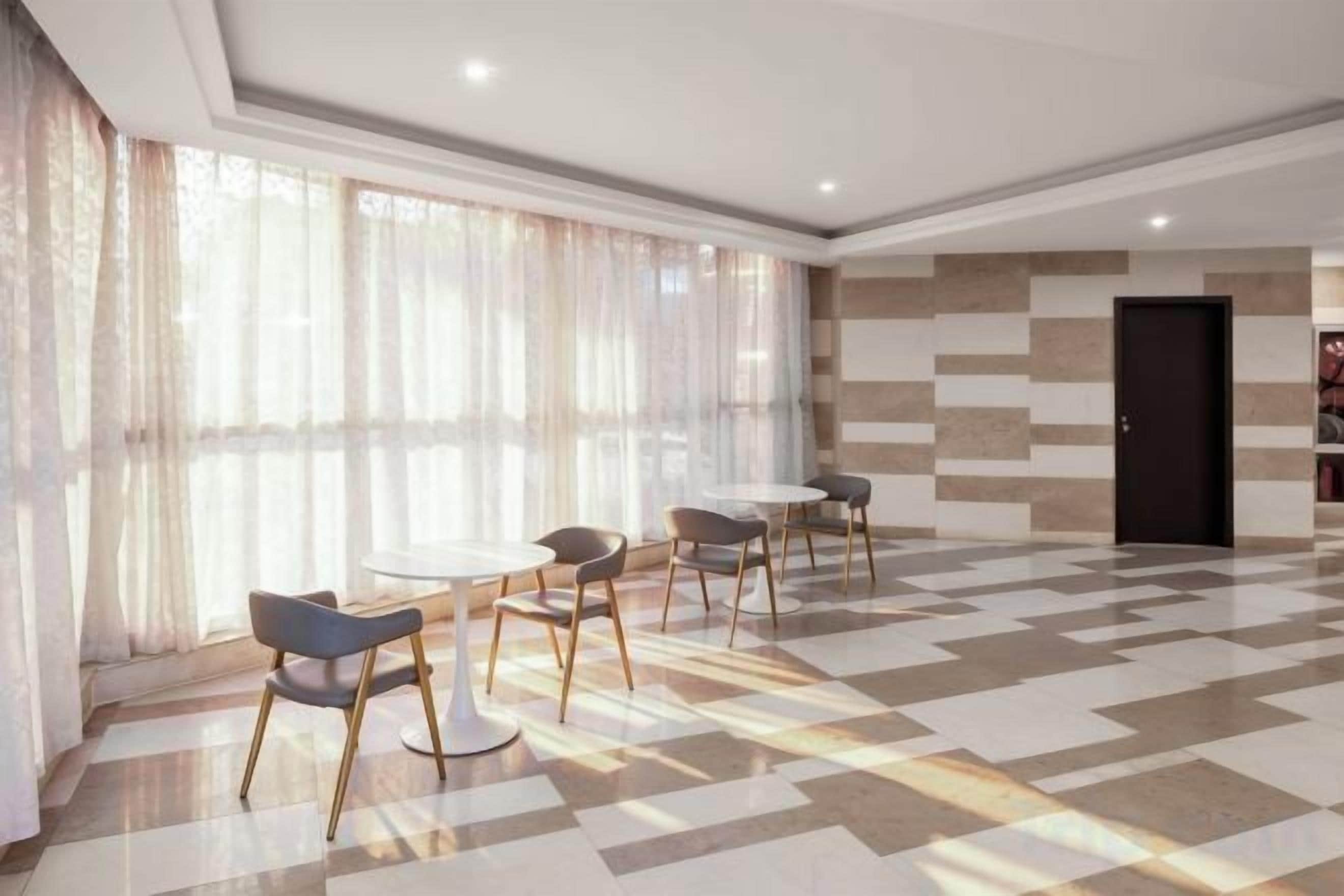 Eine Hotelhalle mit Schachbrettmuster-Boden, einer Sitzecke mit runden Tischen und Stühlen sowie einem großen Fenster mit durchscheinenden Vorhängen.