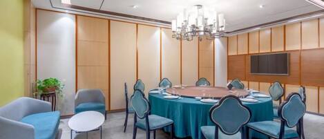 Un salón de conferencias con una mesa redonda, sillas, una araña, un televisor y una planta.