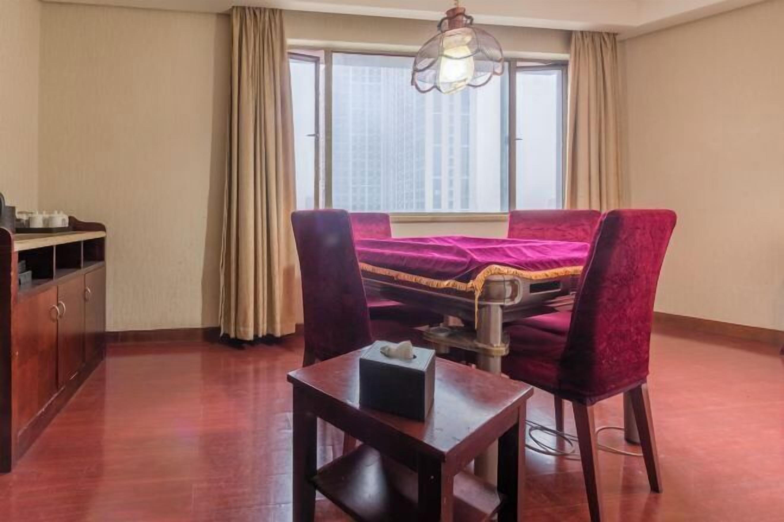 Una habitación de hotel con cama, dos sillas, una mesita, un escritorio, ventana con cortinas y una lámpara colgante.
