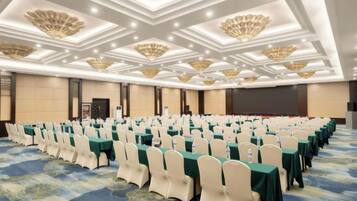 Un salón de conferencias con filas de mesas y sillas, una pantalla grande y un escenario.