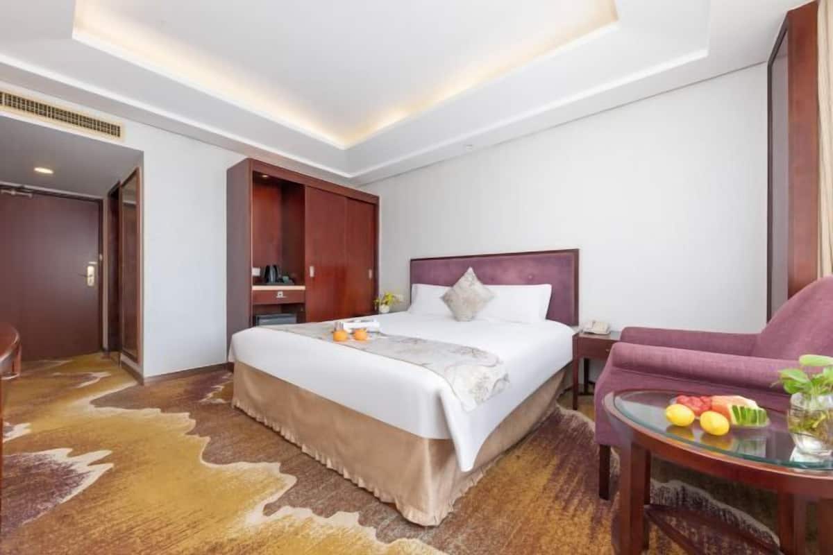 Habitación de hotel con una cama grande, un armario de madera, una mesa de vidrio con frutas y un sillón morado.