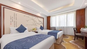 Ein Hotelzimmer mit zwei Betten, einem Schreibtisch, einem Stuhl, einem großen Fenster mit Vorhängen sowie einer Wand mit Kalligraphie.