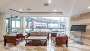 Eine Hotellobby mit einem großzügigen Sitzbereich, einem Flachbildfernseher und großen Fenstern mit Blick auf die Straße und umliegende Gebäude.