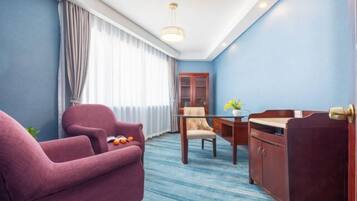 Quarto de hotel com parede azul, escrivaninha de madeira, poltrona roxa, poltrona bege e mesa de vidro.