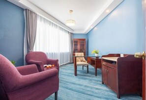 Hotellihuone, jossa on sininen seinä, puinen työpöytä, violetti nojatuoli, beigenvärinen nojatuoli ja lasipöytä.