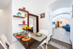 A dining area with a table set for two, a coffee pot, and a fruit bowl. There are white chairs, a wooden shelf with plants, and a view into the living area through an archway. - La Casa Sulla Roccia Trulli Pool (Monopoli)