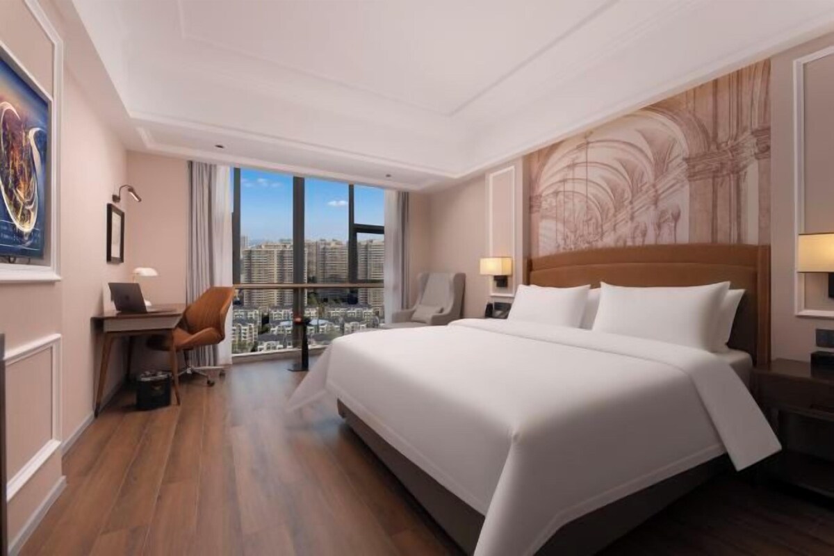 Quarto de hotel com uma cama grande, uma escrivaninha, uma cadeira, uma mesa de cabeceira e vista para a cidade através de amplas janelas.