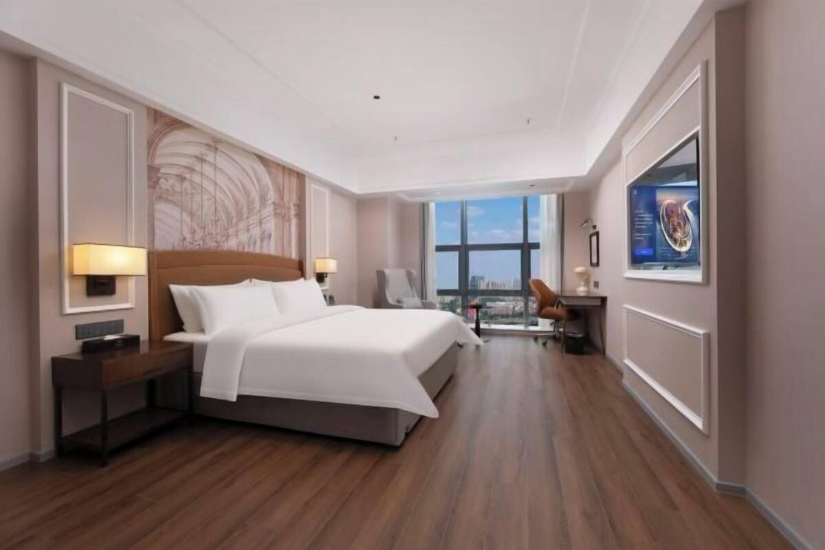 A modern hotel room with a large bed, a desk, a chair, a TV, and a view of the city.