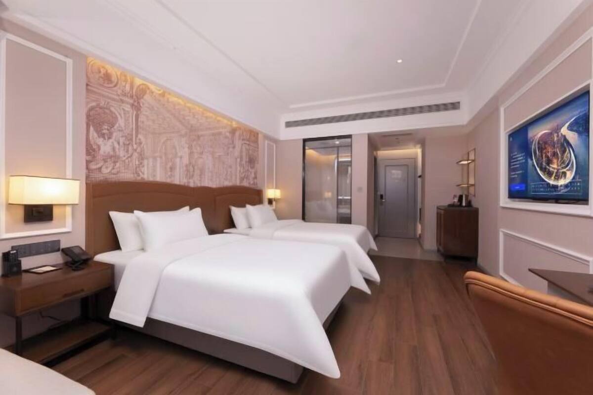 A hotel room with two beds, a nightstand, a desk, a chair, a wall-mounted TV, and a door leading to another room.
