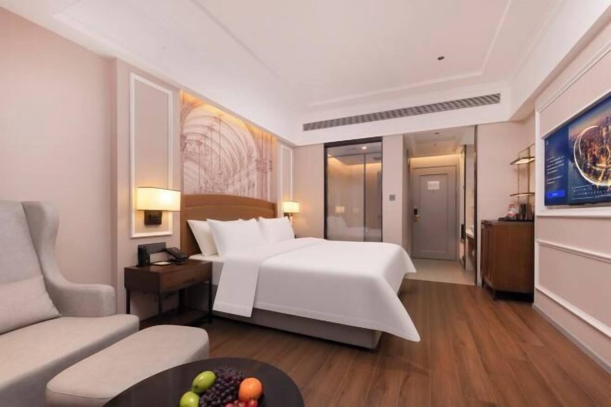 A hotel room with a bed, a desk, a chair, a coffee table with fruit, and a wall with a circular design.