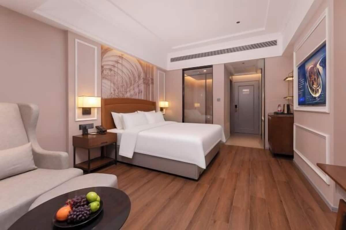 A hotel room with a bed, a desk, a chair, a dining table with fruit, and a painting on the wall.