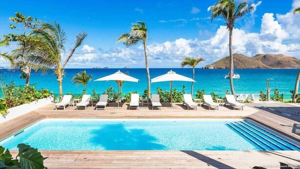A poolside area with lounge chairs and umbrellas, overlooking a clear blue sea and a mountainous backdrop.
