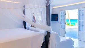 A room with a bed covered by a white canopy, a television on the wall, and a view of the sea through an open door.