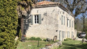 Un edificio de piedra con tejado de tejas, una pared de piedra y un jardín con una carretilla y plantas.