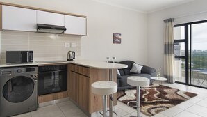 Une cuisine moderne équipée d’un lave-linge, d’un four et d’un four à micro-ondes intégrés. Un petit coin bar avec des tabourets et une vue sur la ville, grâce à de grandes fenêtres, complète l’ensemble.