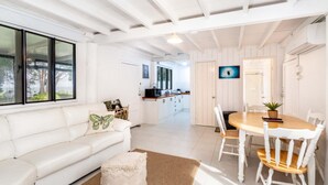 Uma sala de estar moderna, com decoração em tons de branco, incluindo um sofá, uma mesa de jantar com cadeiras e uma cozinha ao fundo.
