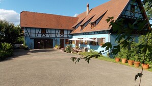 Une maison traditionnelle à colombages, dotée d’un toit à pente, d’un porche couvert avec des sièges d’extérieur et d’un jardin agrémenté de plantes en pot.
