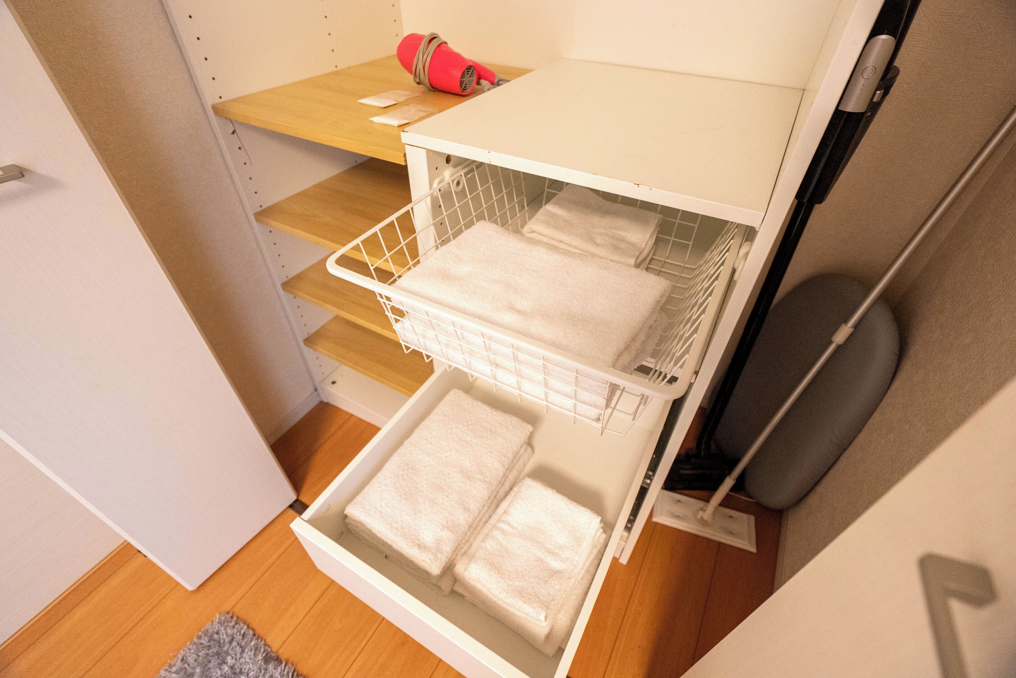 A white laundry basket with folded towels inside, a red hairdryer on a shelf, and a grey cushion on a chair.