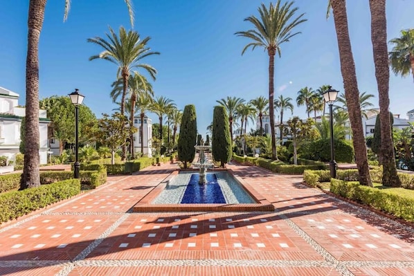 A paved pathway leading to a central fountain, flanked by palm trees and manicured hedges.
