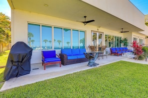 Relax on the patio with a sofa, wicker chair, and dining table, surrounded by a lush lawn and a view of palm trees.