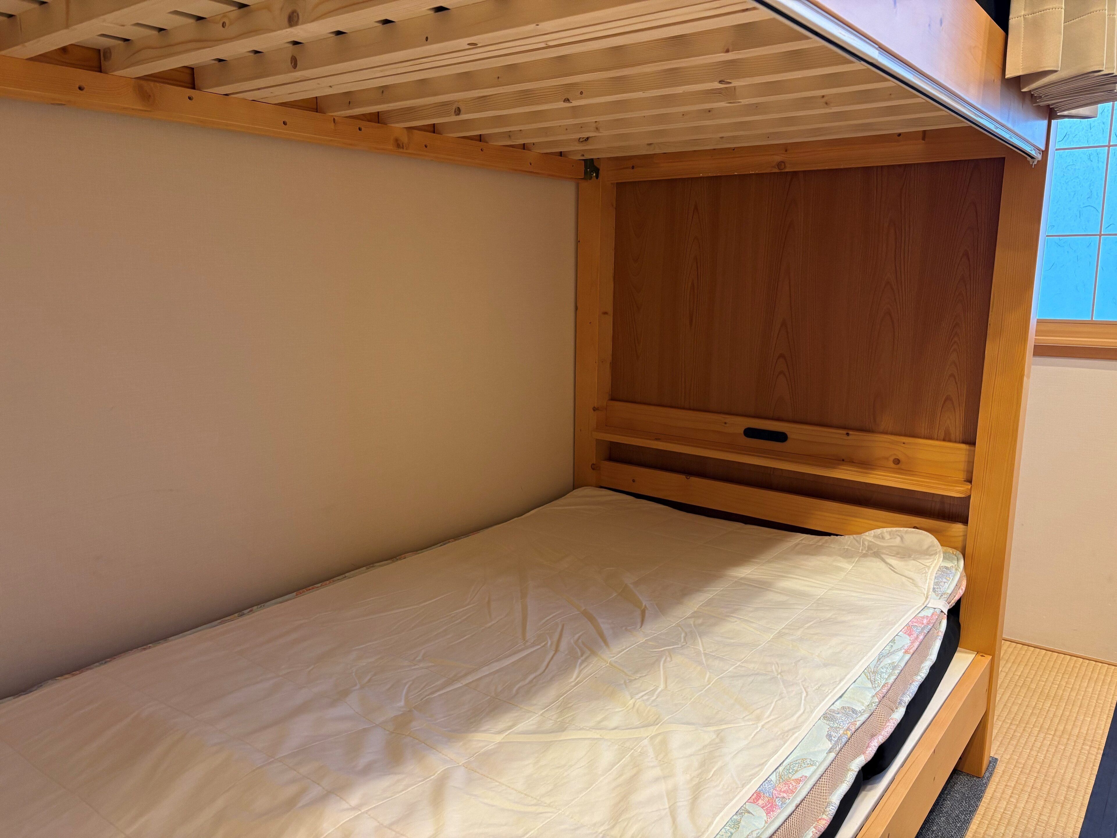 A small, single-bed room with a wooden headboard, a plain wall, and a window with curtains.