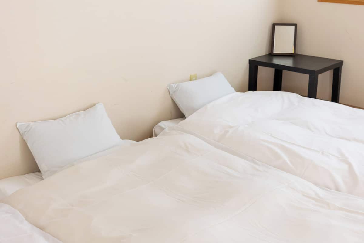 A neatly made bed with white bedding, a small bedside table with a framed picture, and a plain wall.