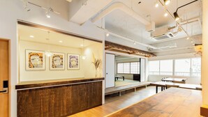 A modern interior with a wooden reception desk, framed artwork, and a seating area with tables and chairs. - Hostel Ogk (Osaka)