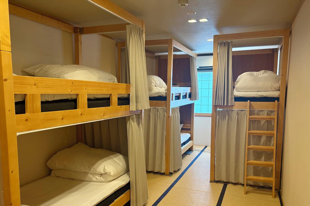 A dormitory-style room with bunk beds, a ladder, and a window with curtains.