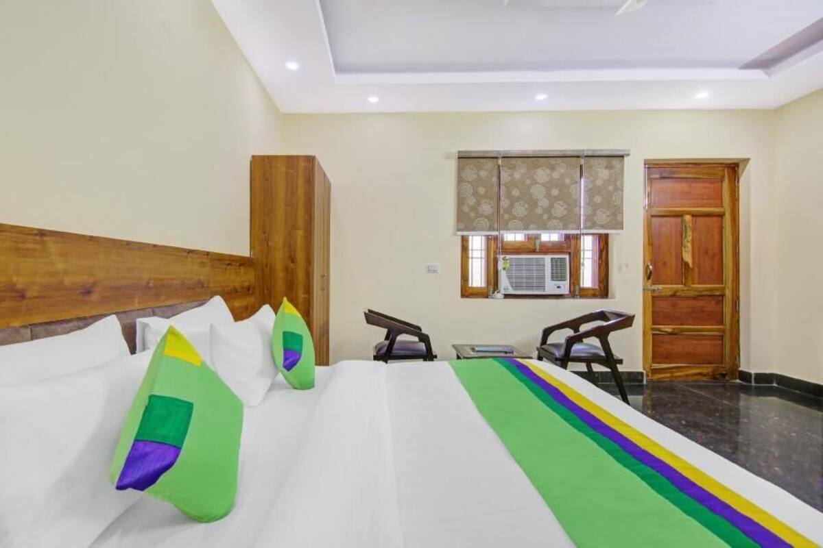 A hotel room with a bed, pillows, a desk, an ironing board, and a window with an air conditioner.