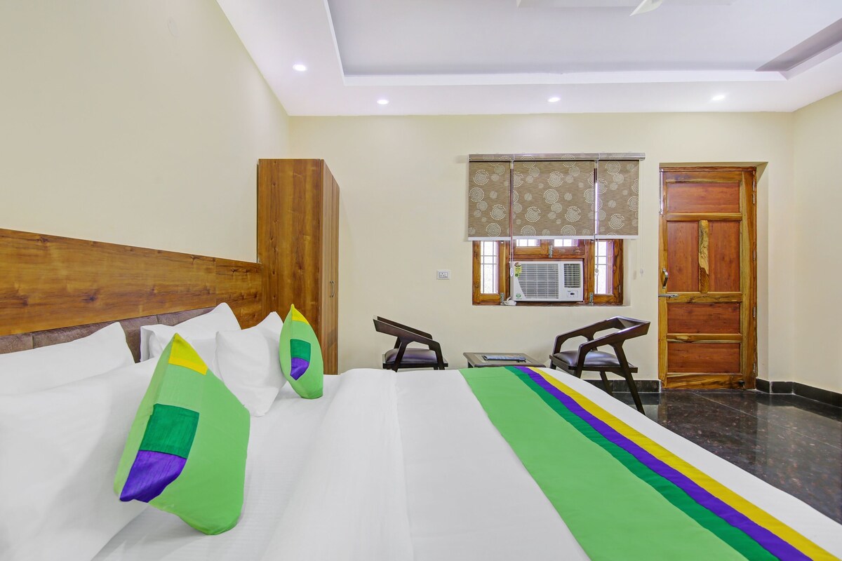 Quarto de hotel com cabeceira de madeira, cama com roupa de cama branca e almofadas coloridas, escrivaninha com cadeira e janela com ar condicionado.