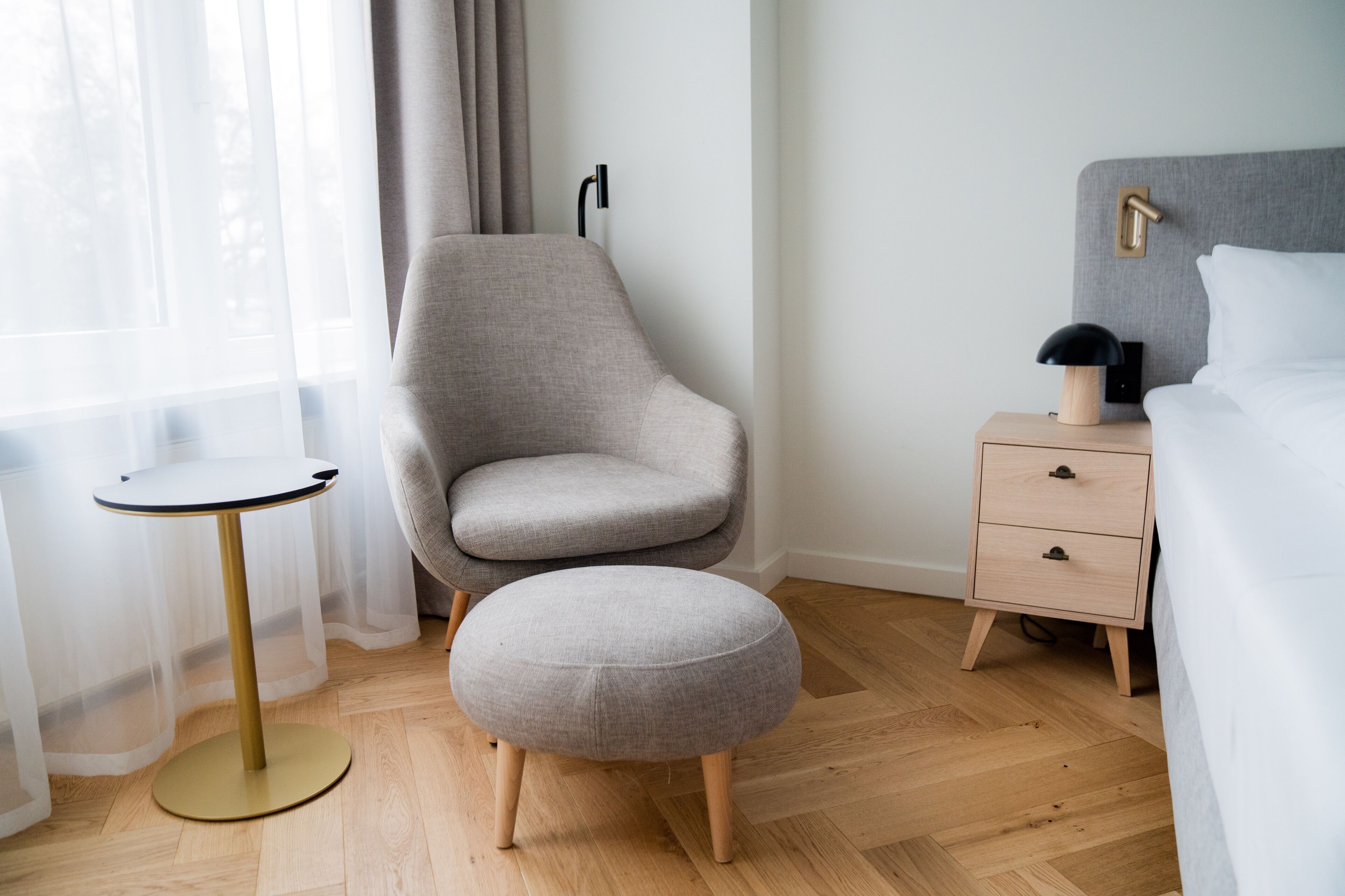 Quarto moderno com poltrona estofada cinza, mesinha redonda, abajur e mesa de cabeceira de madeira.