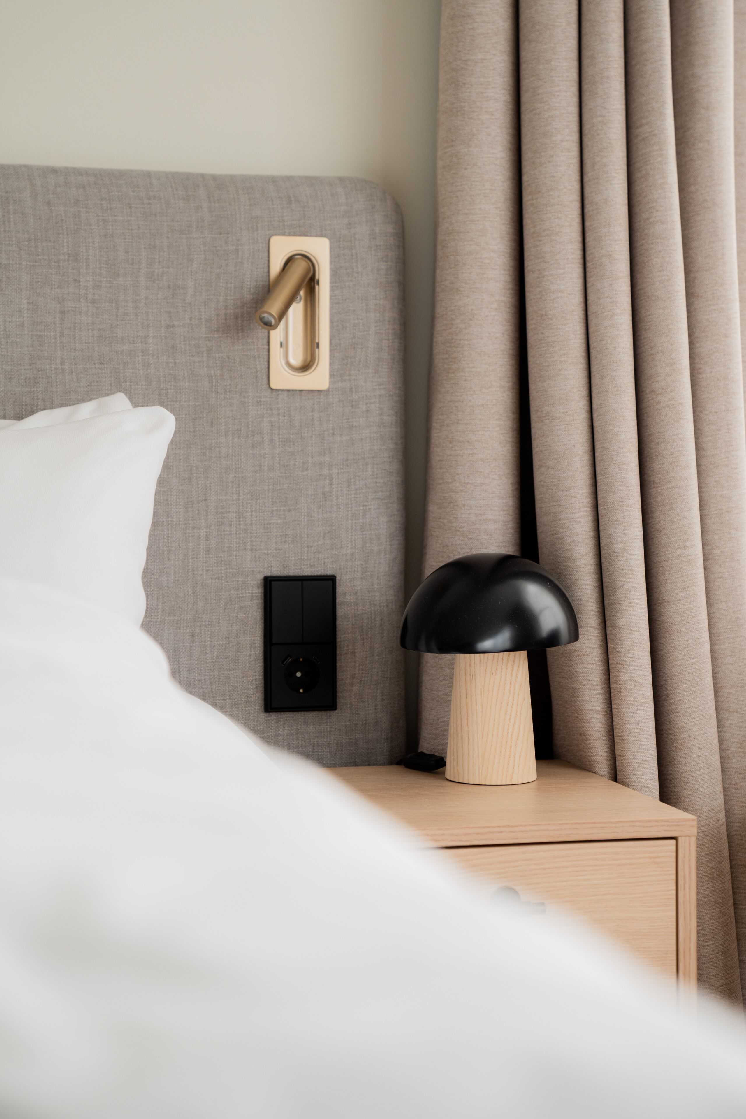 Un lit bien fait, avec un oreiller blanc, une table de chevet en bois surmontée d’une lampe noire, un interrupteur fixé au mur et un rideau.