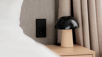 Un lit bien fait, avec un oreiller blanc, une table de chevet en bois surmontée d’une lampe noire, un interrupteur fixé au mur et un rideau.