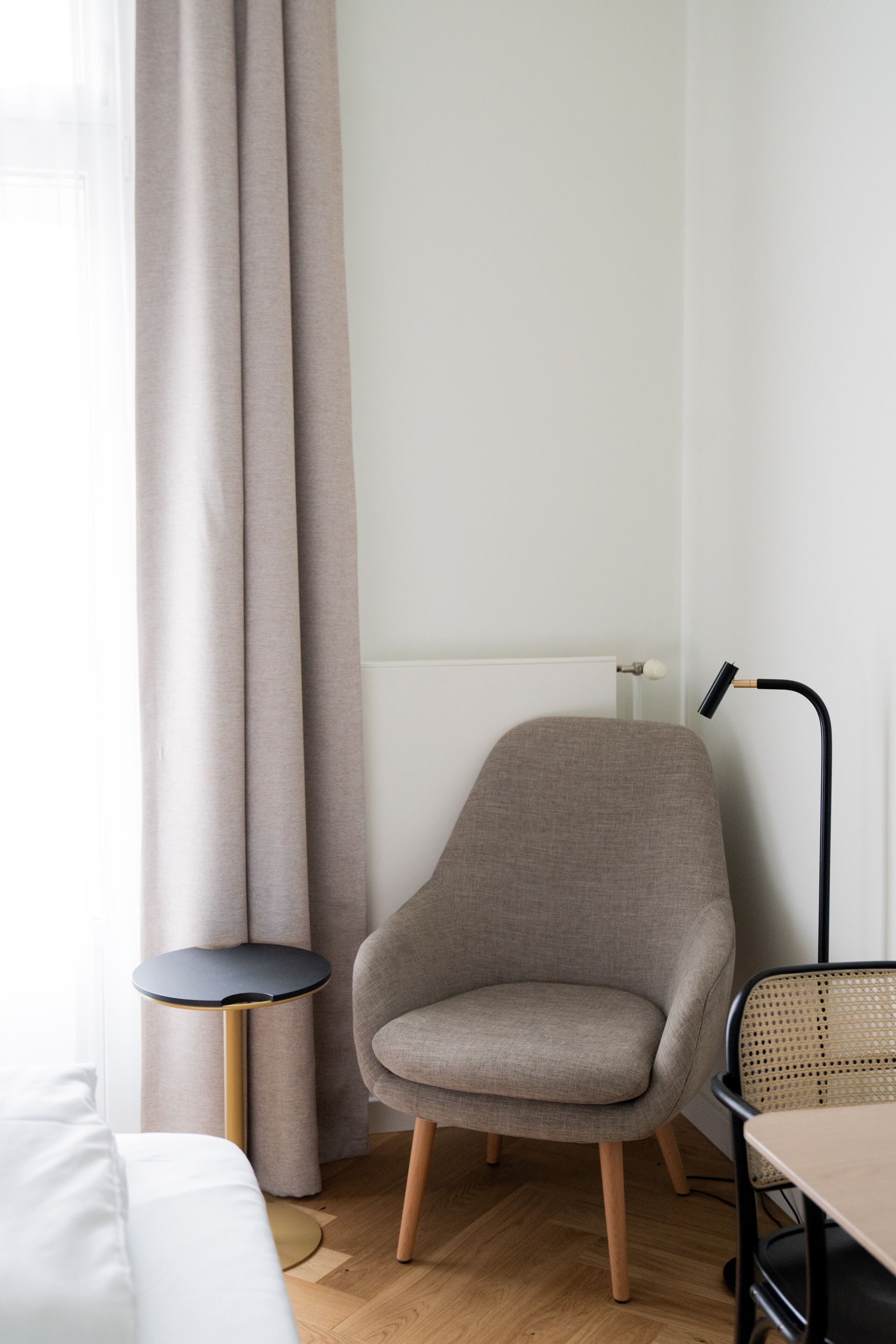 Ein modernes Zimmer mit einem grauen Sessel, einem kleinen runden Tisch, einer Lampe und einem Bett mit weißer Bettwäsche.