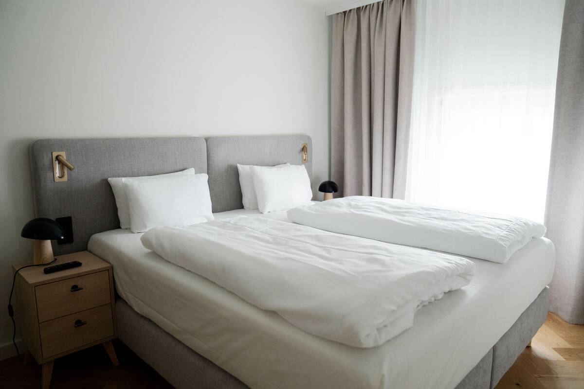 A neatly made bed with white linens, a bedside table with a lamp, and a window with curtains.