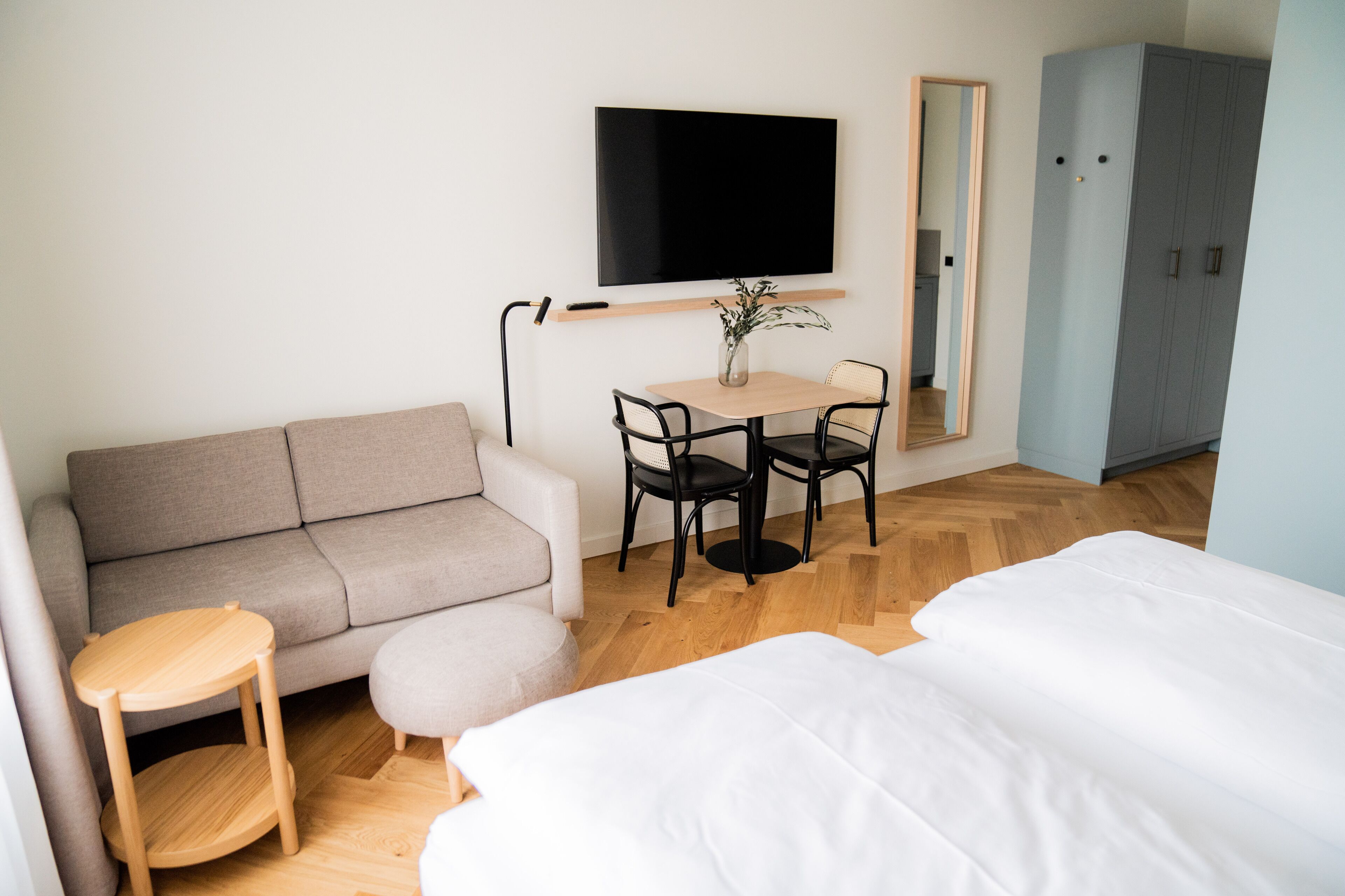 A modern hotel room with a sofa, a small table with chairs, a TV mounted on the wall, and a wardrobe.