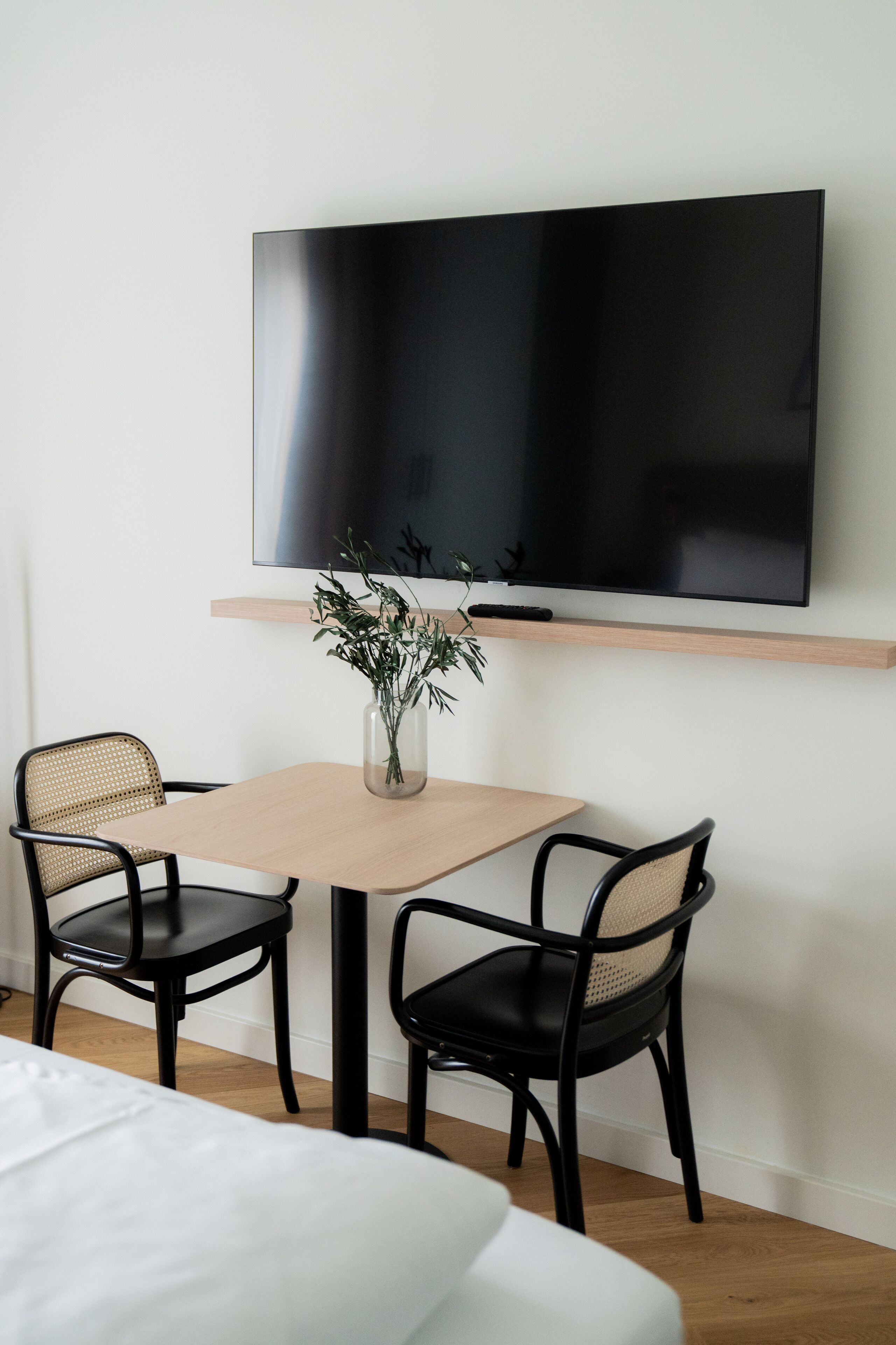 Une chambre d’hôtel moderne avec un téléviseur à écran plat fixé au mur, une table en bois avec deux chaises noires, un vase avec des plantes vertes et un lit avec une literie blanche.
