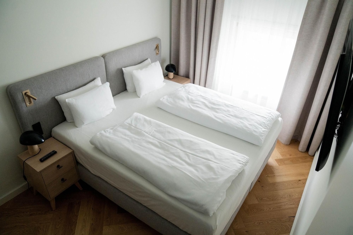 A neatly made double bed with white linens, a bedside table with a lamp, and a nightstand with a remote control.